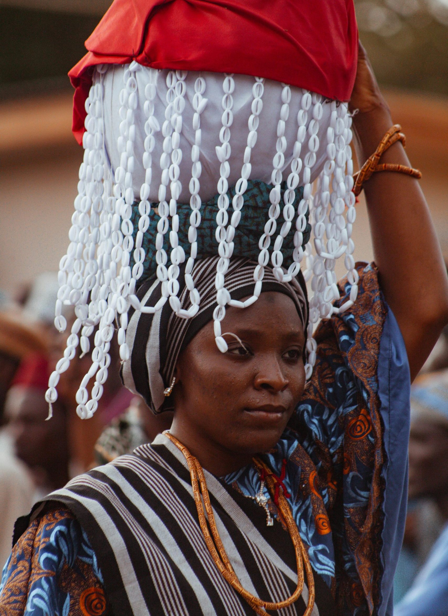 A woman in traditional attire carrying ornate headwear during an outdoor African cultural ceremony.